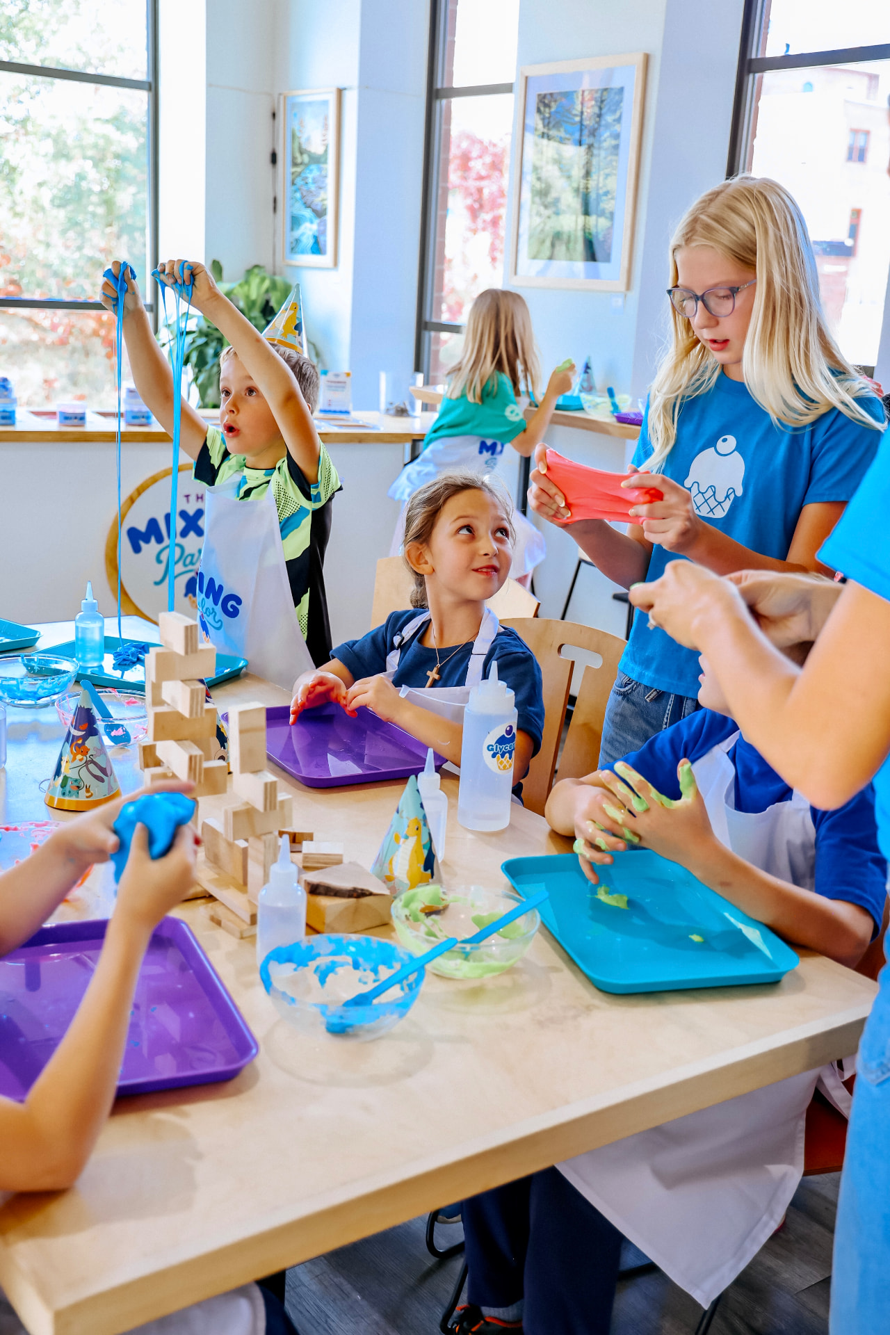 Kids stretching blue and pink slime over a workshop table while an adult helps a child mix a batch