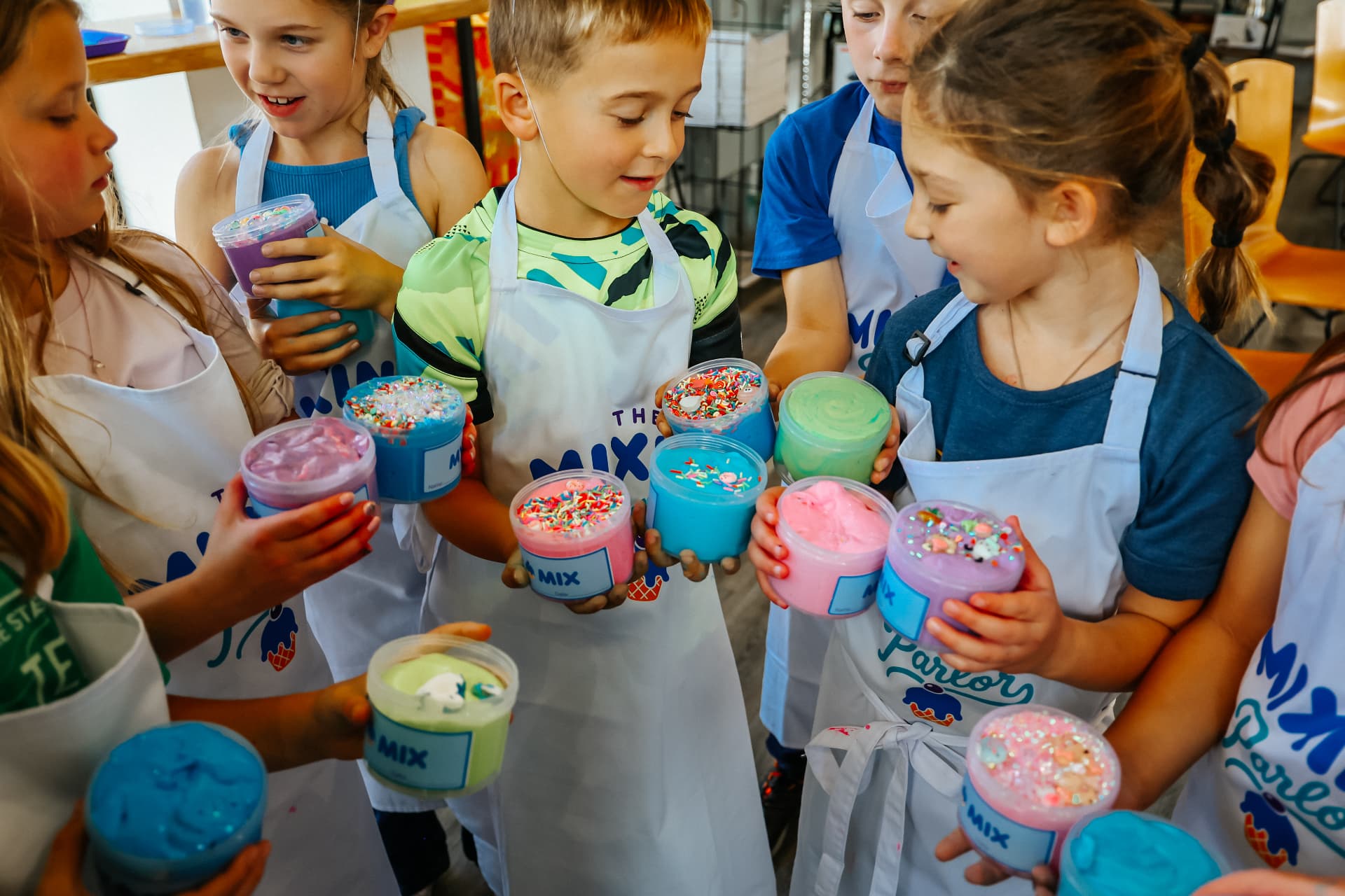 A group of smiling children in white Mixing Parlor aprons holding up tubs of colorful slime topped with sprinkles